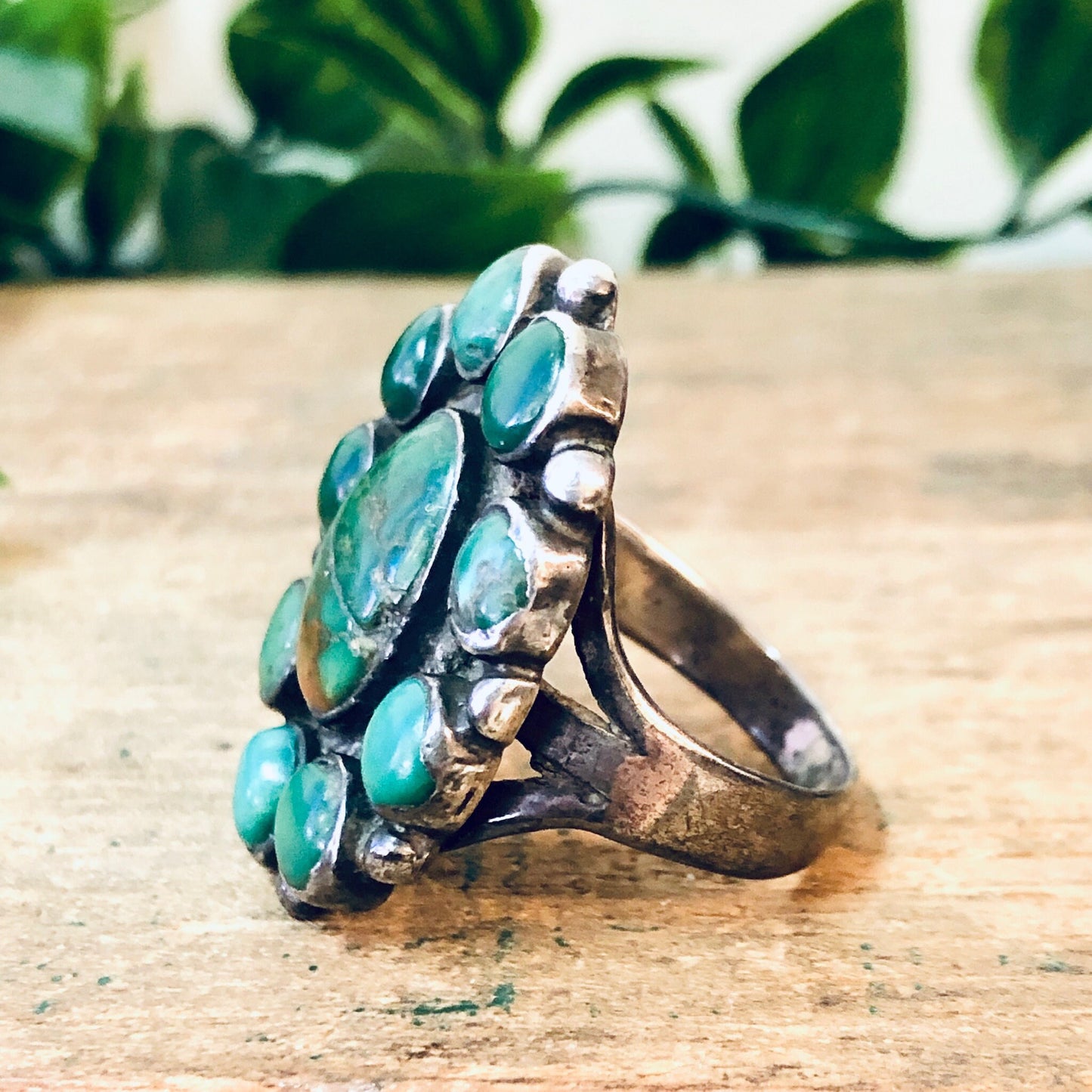 Large sterling silver ring with an oversized turquoise gemstone cluster in shades of teal green, displayed on a wooden surface with foliage in the background, bohemian vintage southwestern statement jewelry.