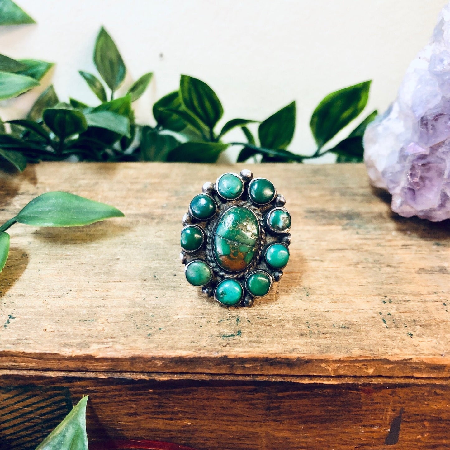 Vintage silver turquoise ring with green turquoise cabochon stone surrounded by smaller turquoise accents, set on wooden surface with leaves and amethyst crystal, bohemian southwestern statement jewelry.