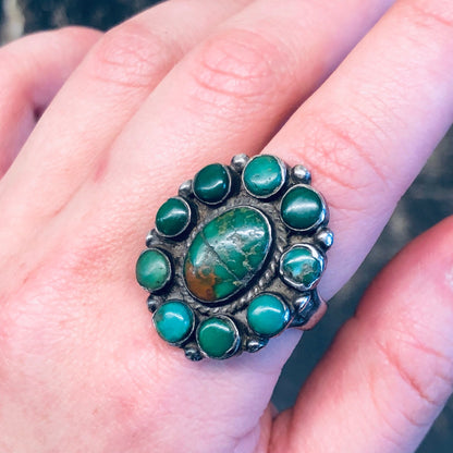 Large silver vintage-style ring with a prominent round green turquoise stone in the center surrounded by smaller round turquoise accents, held in a woman's fingers showing the bohemian southwestern statement jewelry piece