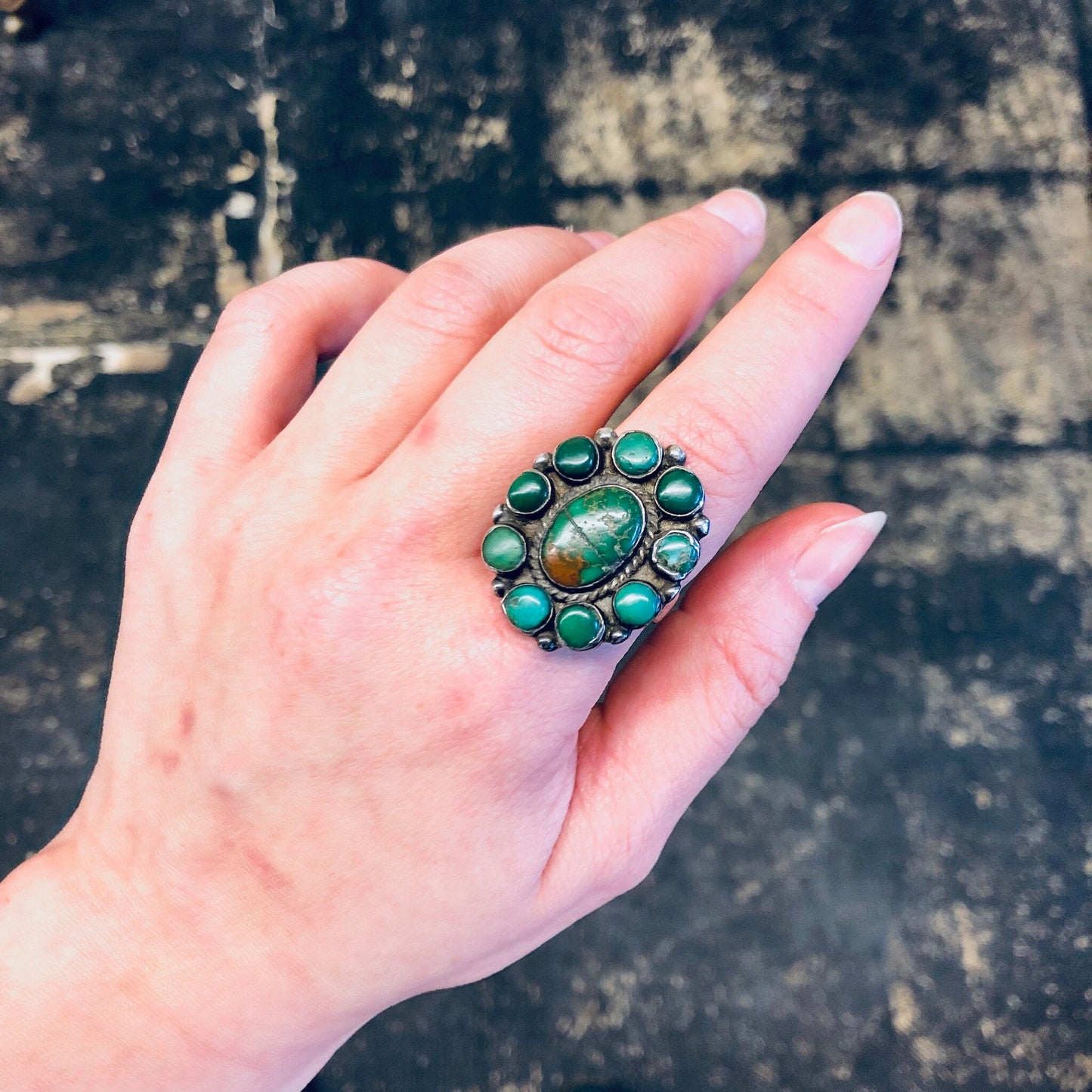 Large turquoise ring with green stones set in vintage silver on a hand, against a grungy background. Bohemian southwestern statement jewelry piece.