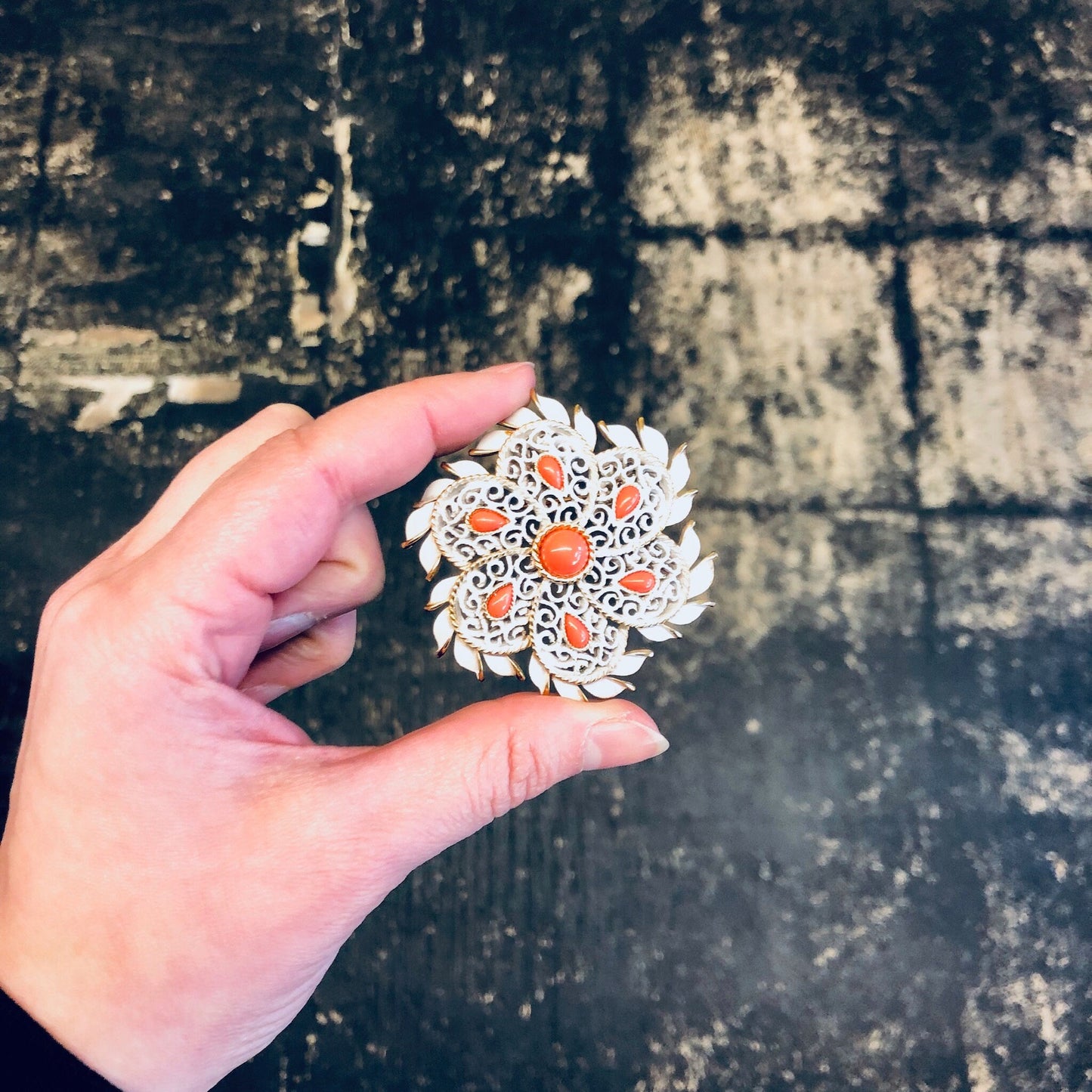 Vintage Trifari white enamel floral brooch with orange accents and gold tone, held in hand against weathered concrete background