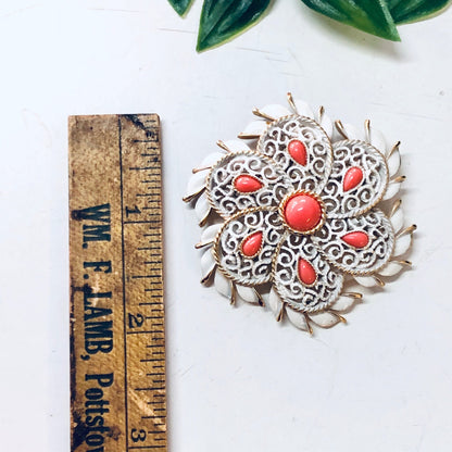 Vintage Trifari white brooch with orange accents and gold-toned filigree design, displayed next to a wooden ruler on a white background with green leaves above.