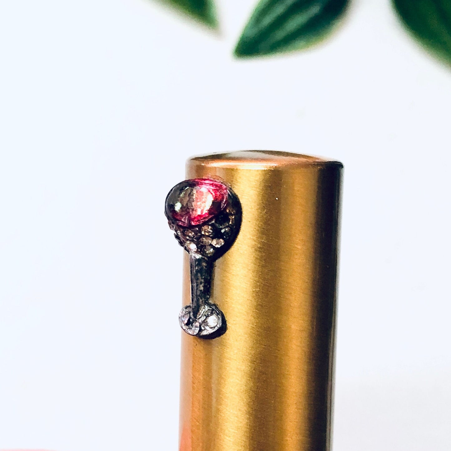 Vintage gold-toned rhinestone compact lipstick tube with red jewel on top, sitting against a white background with green leaves in the foreground, luxury makeup accessory