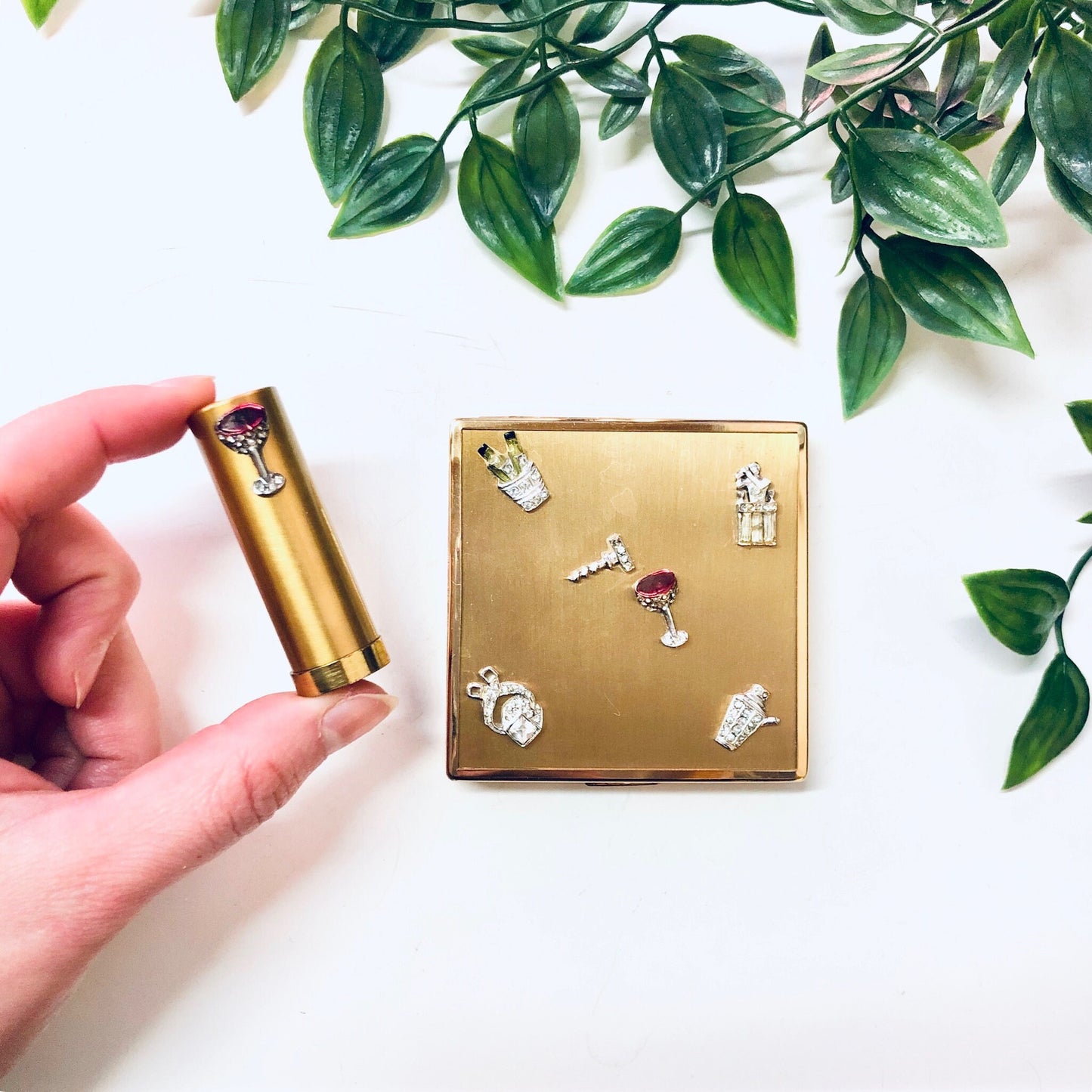 Vintage gold toned compact with rhinestone martini design next to lipstick tube, surrounded by green leaves on white background