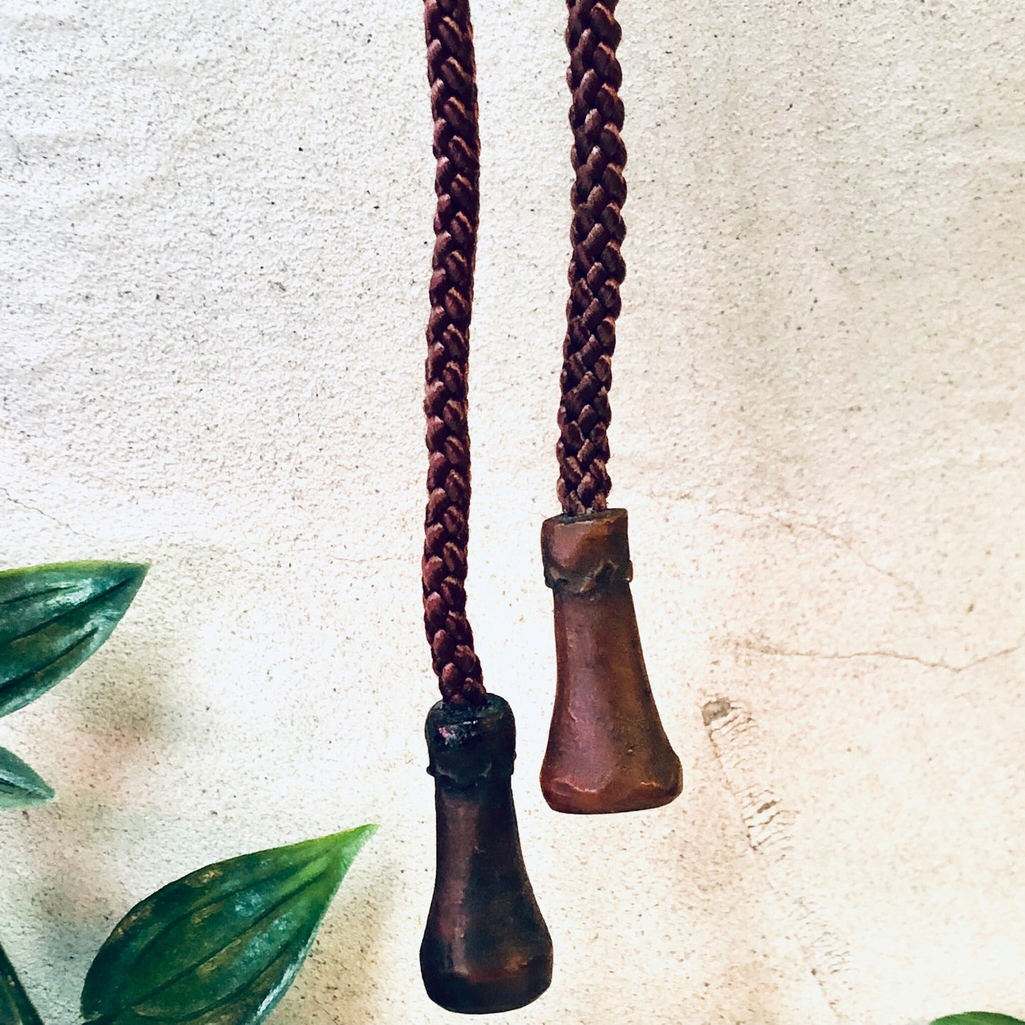 Vintage Japanese bolo tie with carved wooden face pendants and braided rope, alongside green leaves on textured background