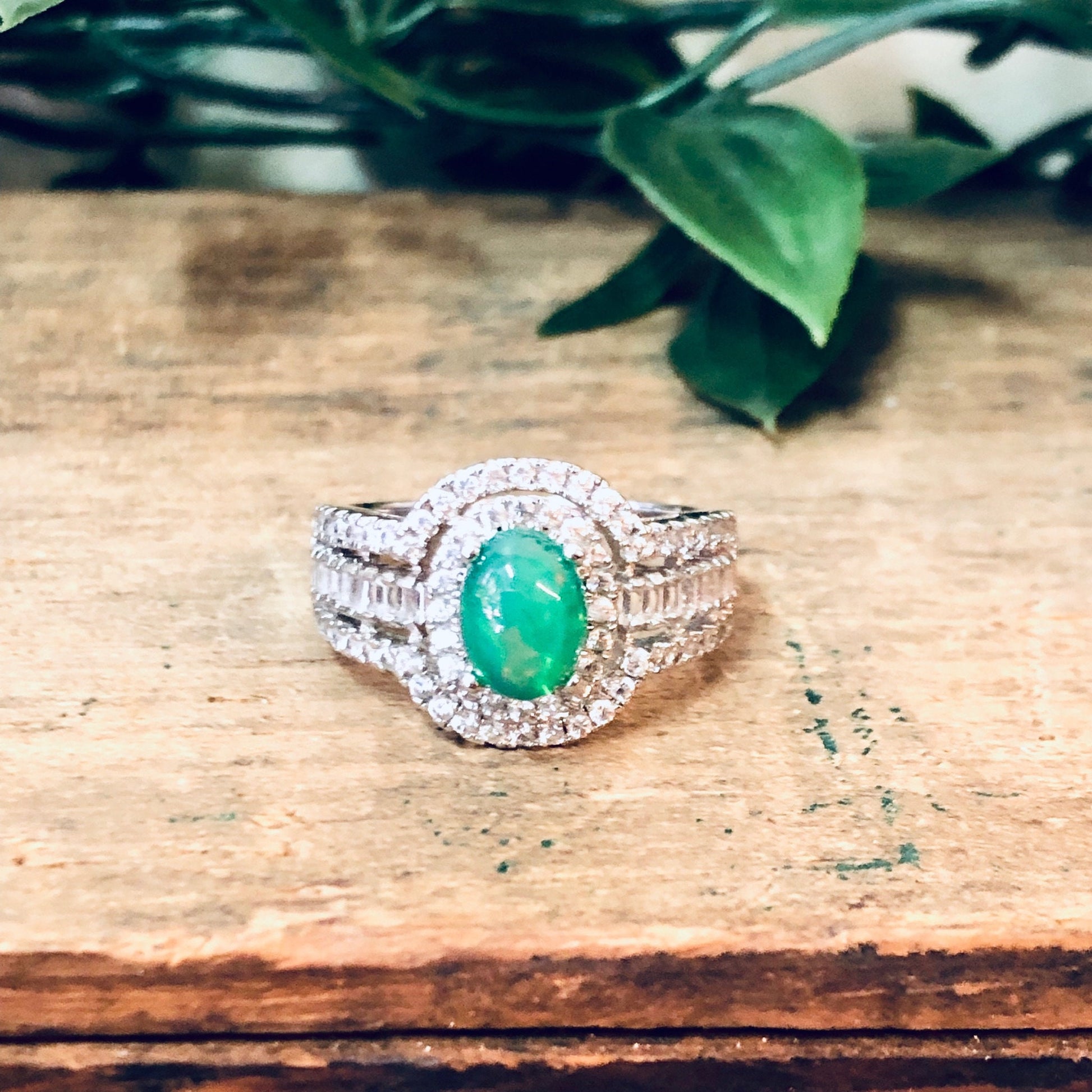 Vintage silver ring with green and opalescent stones surrounded by faux diamonds, photographed on a wooden surface with green foliage in the background