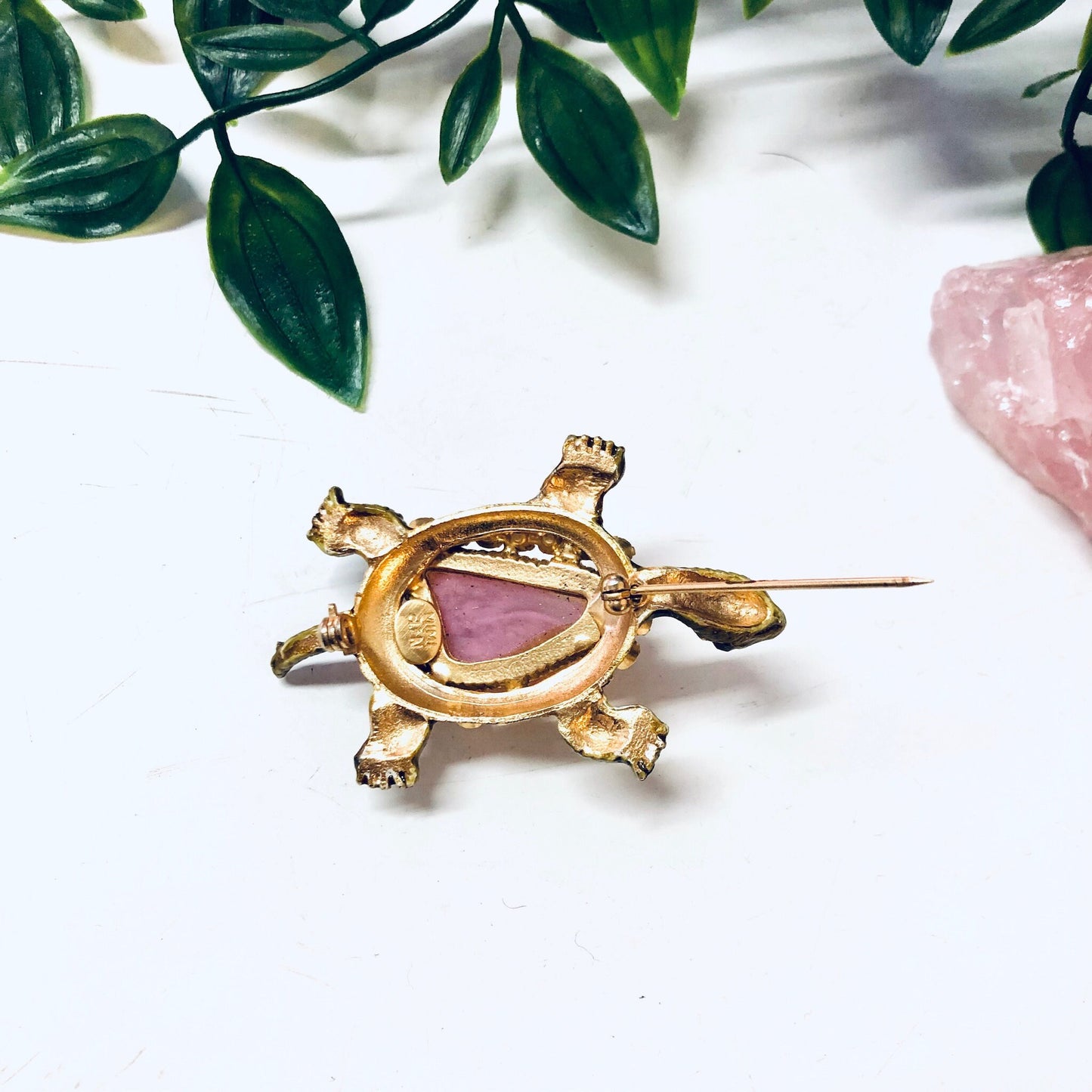 Vintage gold-toned turtle brooch with pink gemstone shell, surrounded by green leaves on a white background.