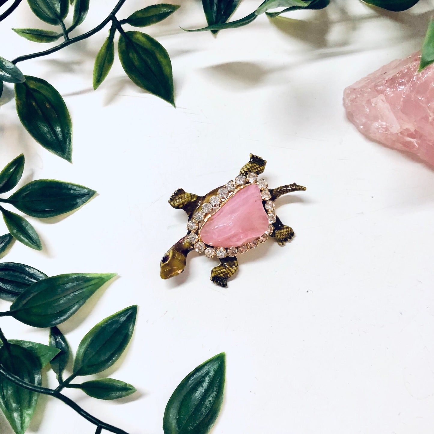 Vintage gold-toned turtle brooch pin with pink gemstone shell and rhinestone accents, surrounded by green leaves, on a white background.