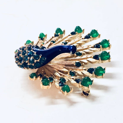 Vintage gold-toned Boucher peacock brooch pin with blue gemstones and green beads, resembling a majestic peacock with its tail feathers fanned out, glimmering in the light.