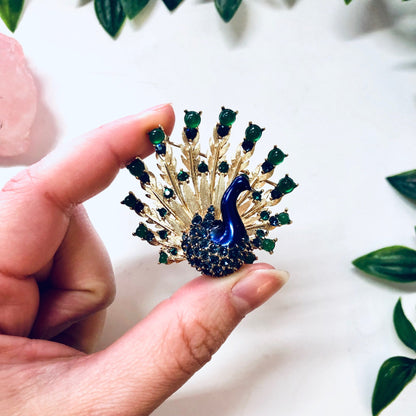 Vintage gold-toned Boucher peacock brooch pin with blue gemstones and green beads, held in hand against white background with green leaves.