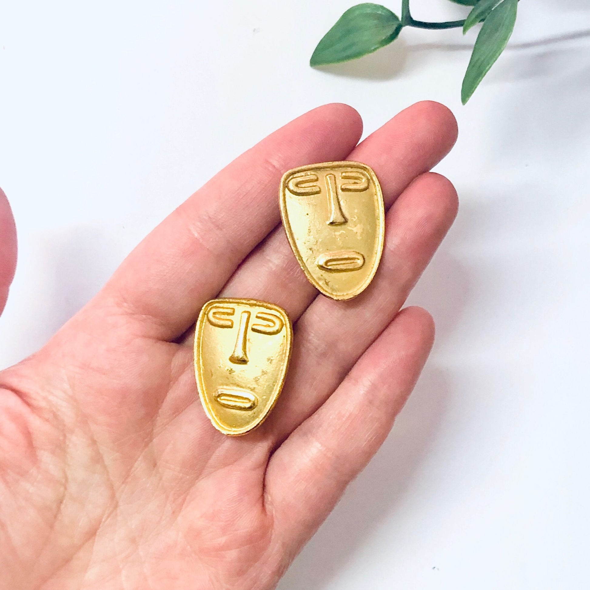 Vintage gold-toned abstract face clip-on earrings held in palm of hand against white background with green leaves