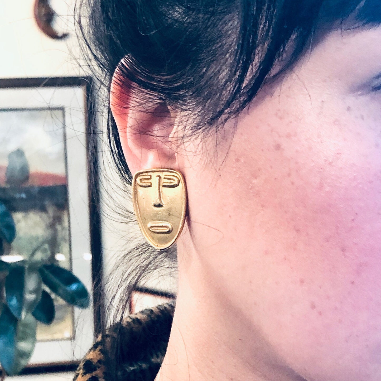 Gold abstract face clip-on earrings worn on ear, with black hair visible in background.