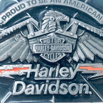 Vintage Harley Davidson belt buckle with eagle design, featuring "Proud To Be An American" text and red stripe accents on a weathered silver background, collectible biker accessory.