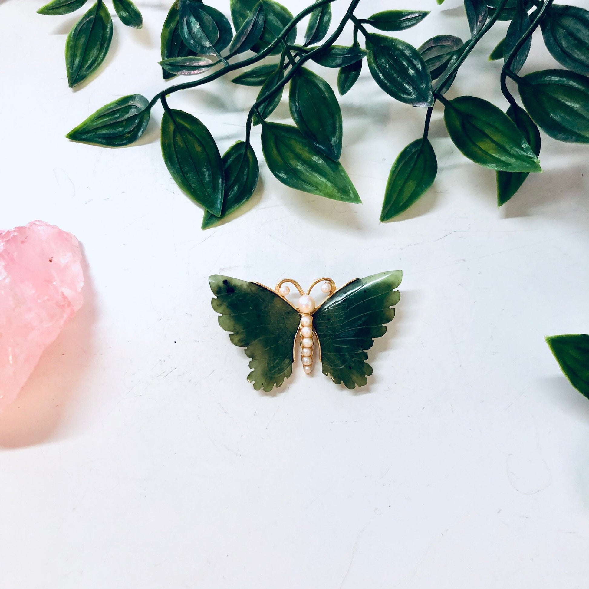 Vintage gold-toned butterfly brooch with green jade wings and faux pearl accents, surrounded by lush green leaves, on a white background with a pink crystal.