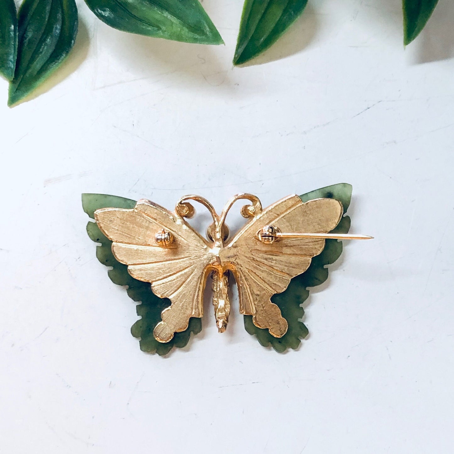 Vintage gold-toned butterfly brooch with faux pearl body and green jade wings, surrounded by leaves on a white background