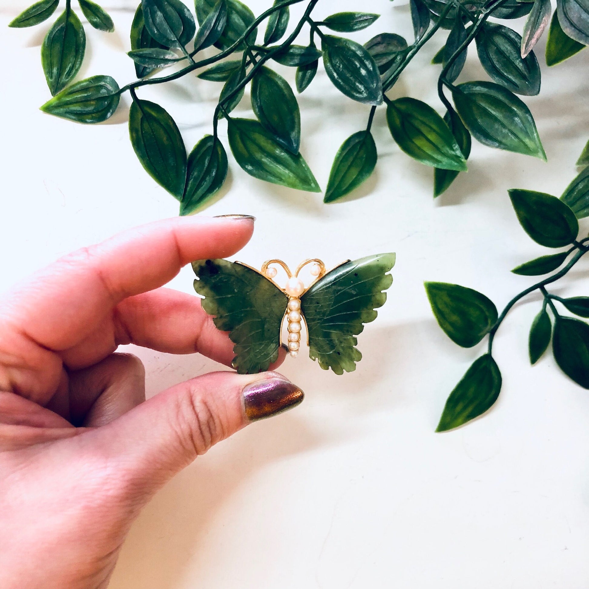 Vintage gold-toned butterfly brooch with green jade wings and faux pearl body held in hand with green leaves in background