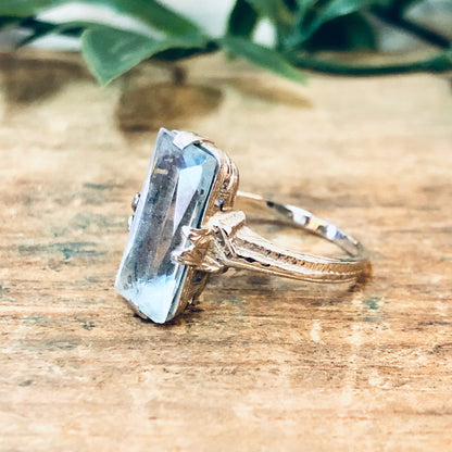 Vintage 18K white gold aquamarine ring with light blue center stone and intricate detailing, photographed on wooden surface with leaves in background