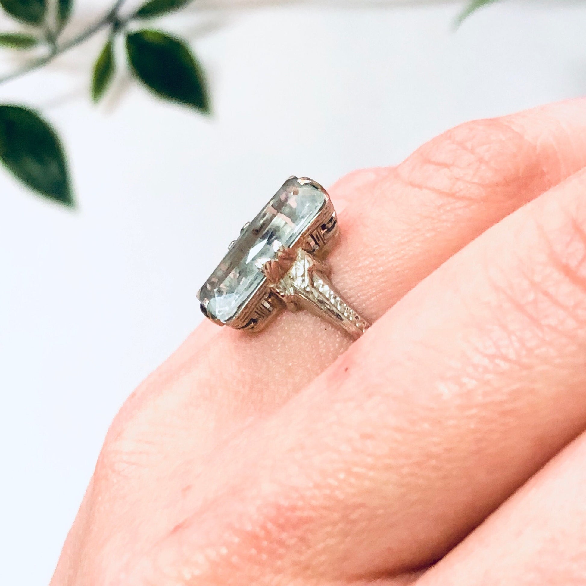 Vintage 18K white gold aquamarine ring with light blue gemstone, pictured on a hand with leaves in the background.