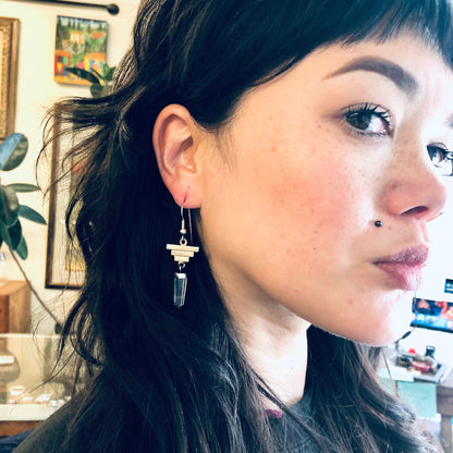 The image shows a close-up of a woman with long dark hair wearing dangle earrings made of clear quartz crystals and silver. The earrings have a pyramid-shaped design and hang from the woman's ear. The woman appears to be looking slightly to the side and has a thoughtful expression on her face.