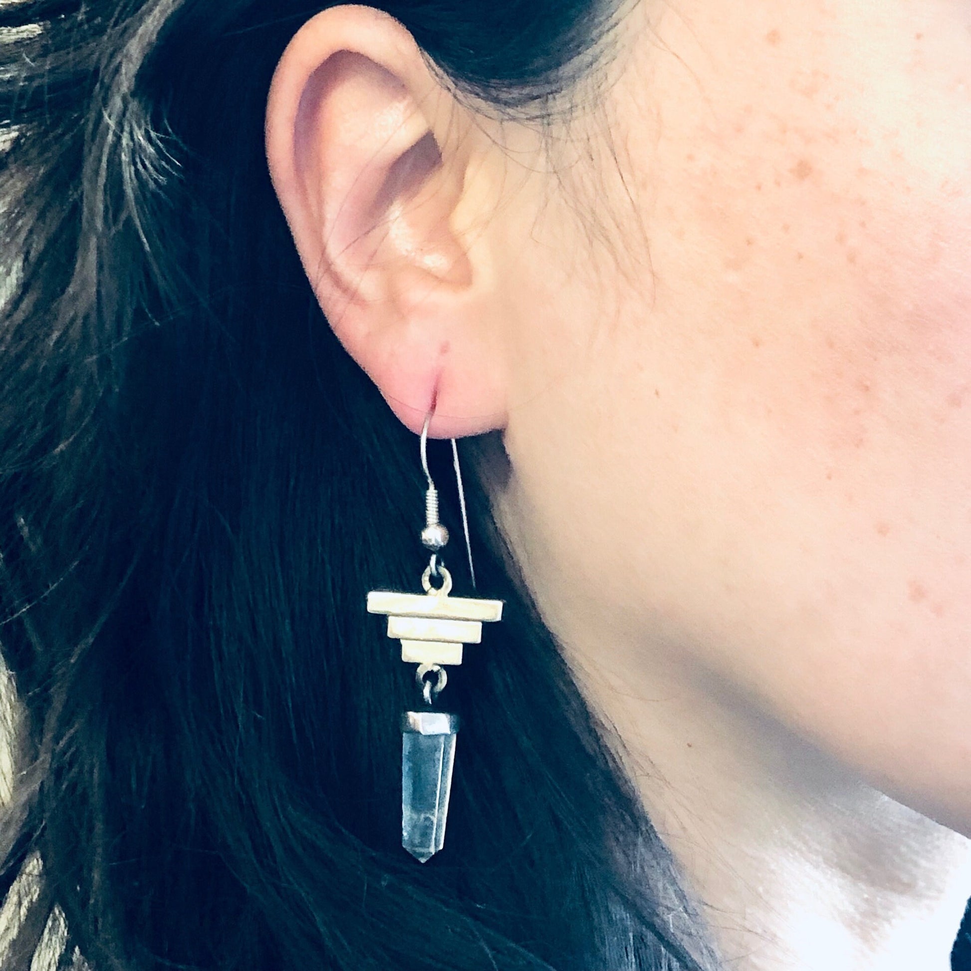 The image shows a close-up of a woman's ear wearing a dangly silver earring. The earring features a small quartz crystal point hanging from a silver geometric pyramid shape. The woman has long dark hair and her ear is shown in profile against a light background.