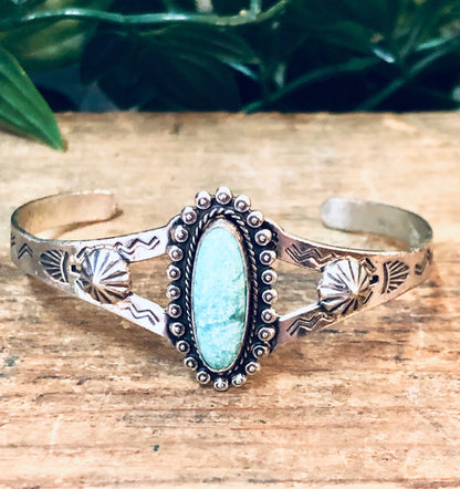 Vintage silver cuff bracelet with turquoise stone and stamped floral design on weathered wooden surface with greenery in background