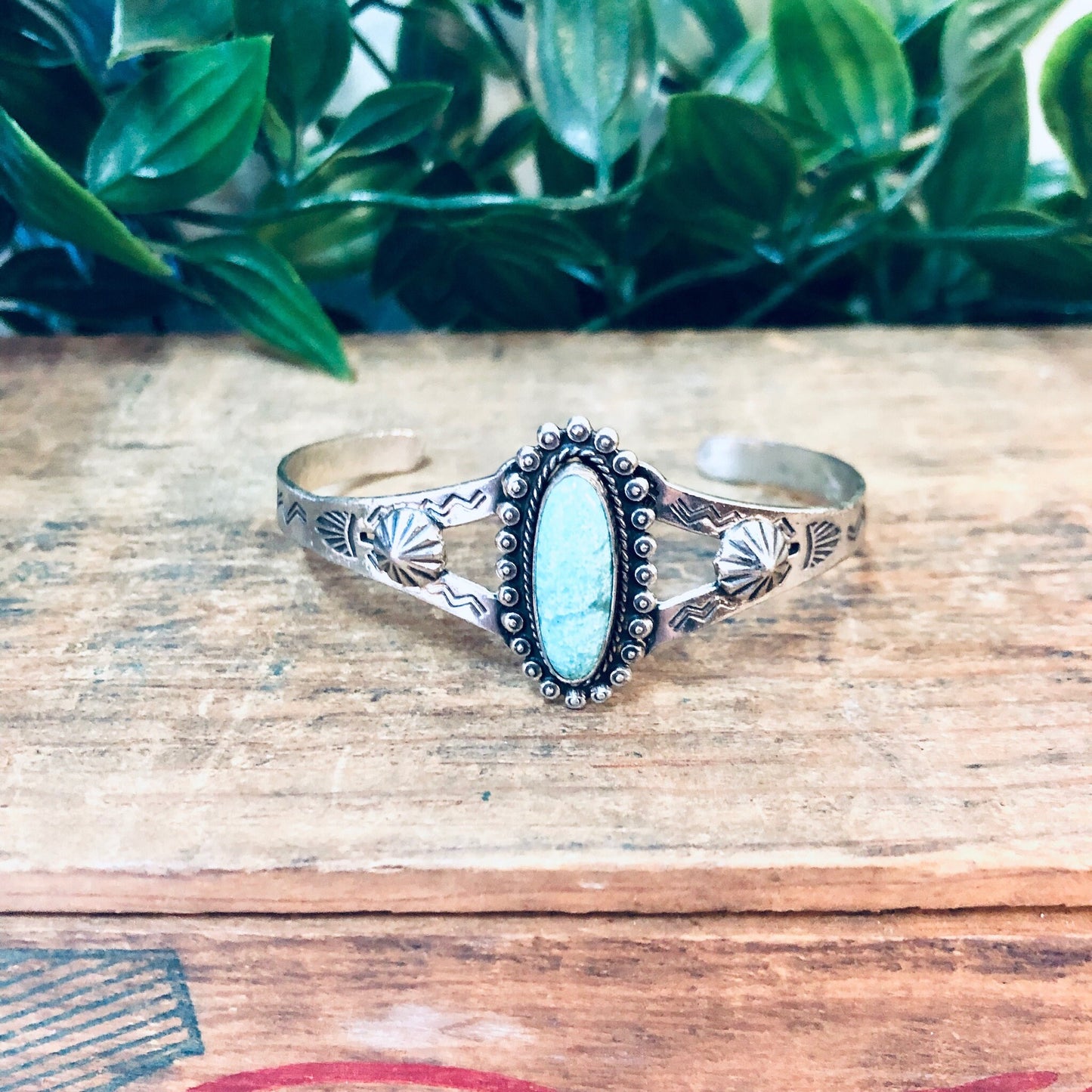 Vintage silver cuff bracelet with oval turquoise stone centerpiece and stamped decorative design, placed on a weathered wooden surface with green foliage in the background.