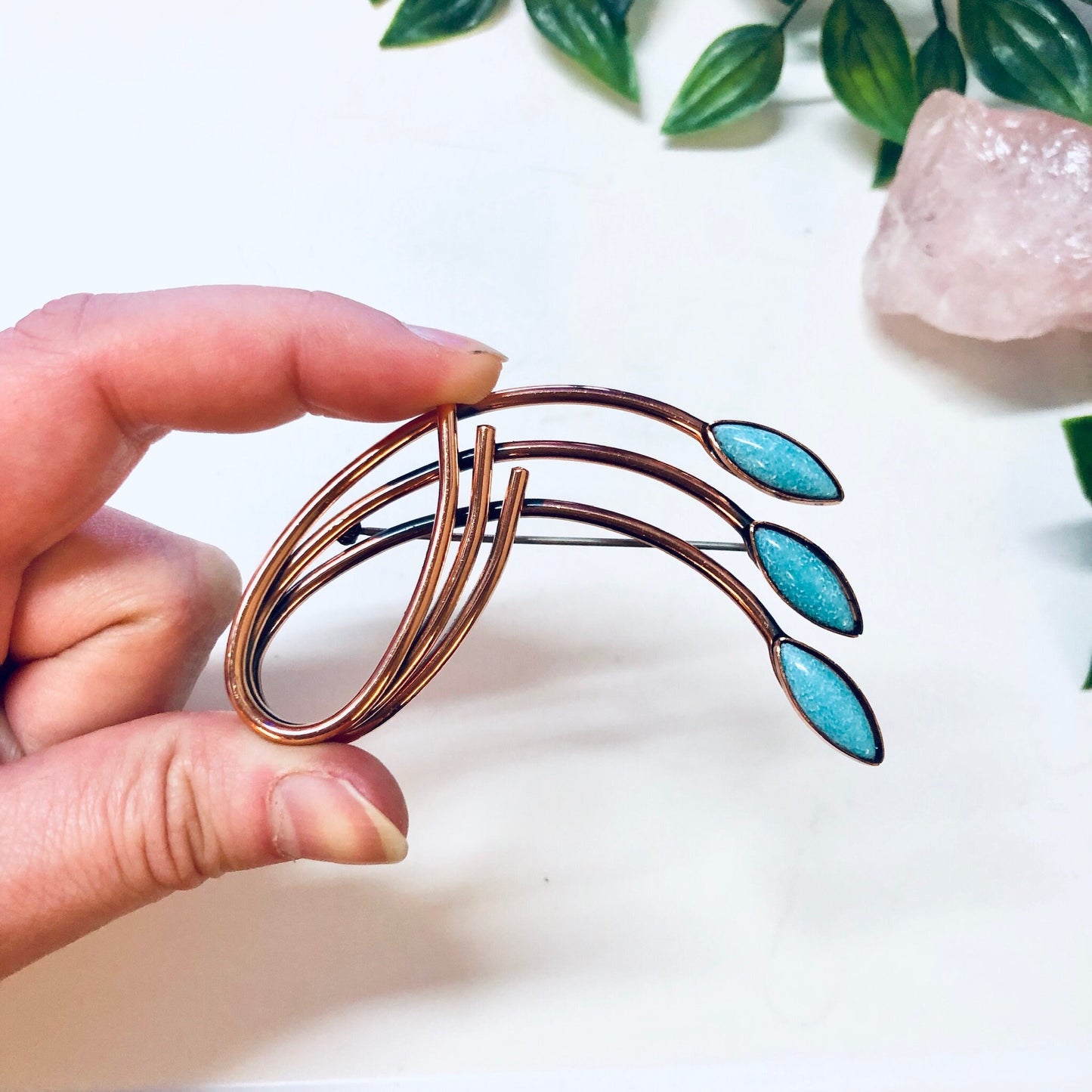 Copper and turquoise vintage Matisse Renoir brooch pin held in hand with decorative leaves and rose quartz crystal in background.