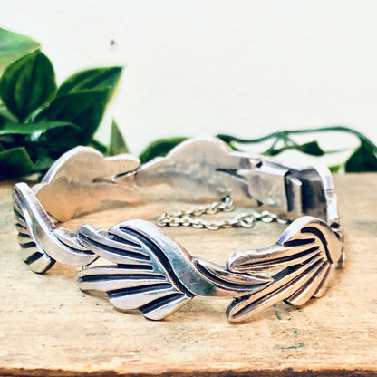 Vintage Taxco silver link bracelet with modernist leaf design, crafted by renowned Mexican silversmith J. Flores, displayed on wooden surface with green foliage in background.