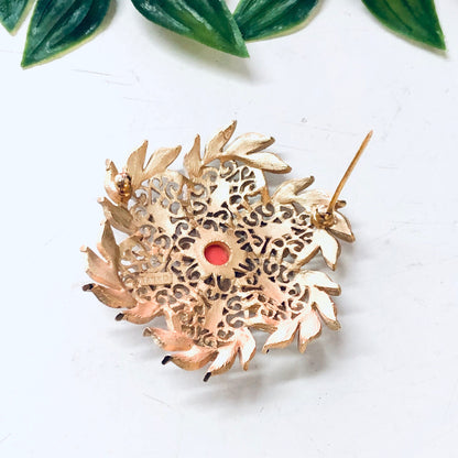 Vintage gold-tone Trifari brooch featuring intricate white enamel floral design with orange accents, displayed on white surface with green leaves in background.