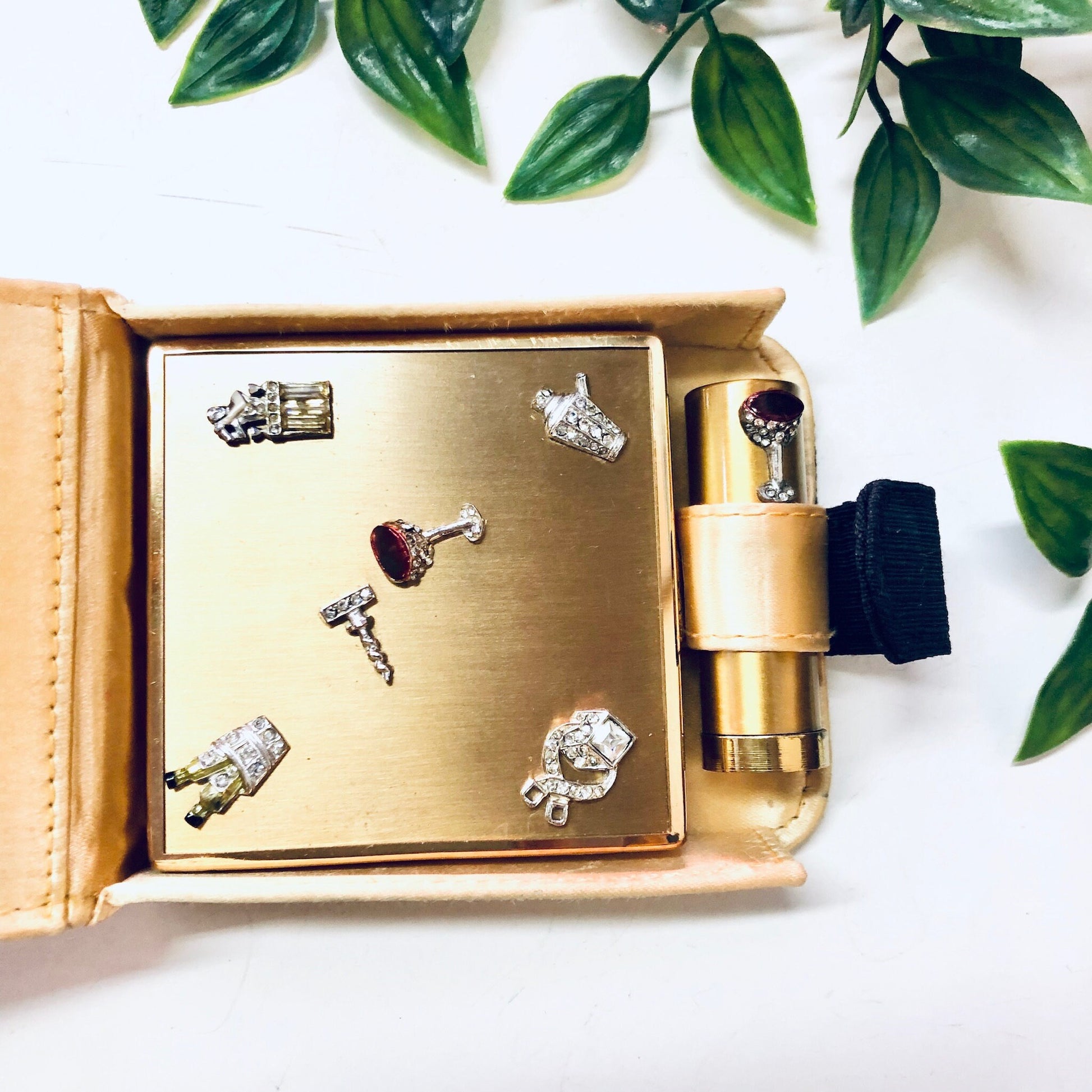 Vintage gold-toned Ciner compact with rhinestone martini glass and lipstick charms, displayed with greenery on white background