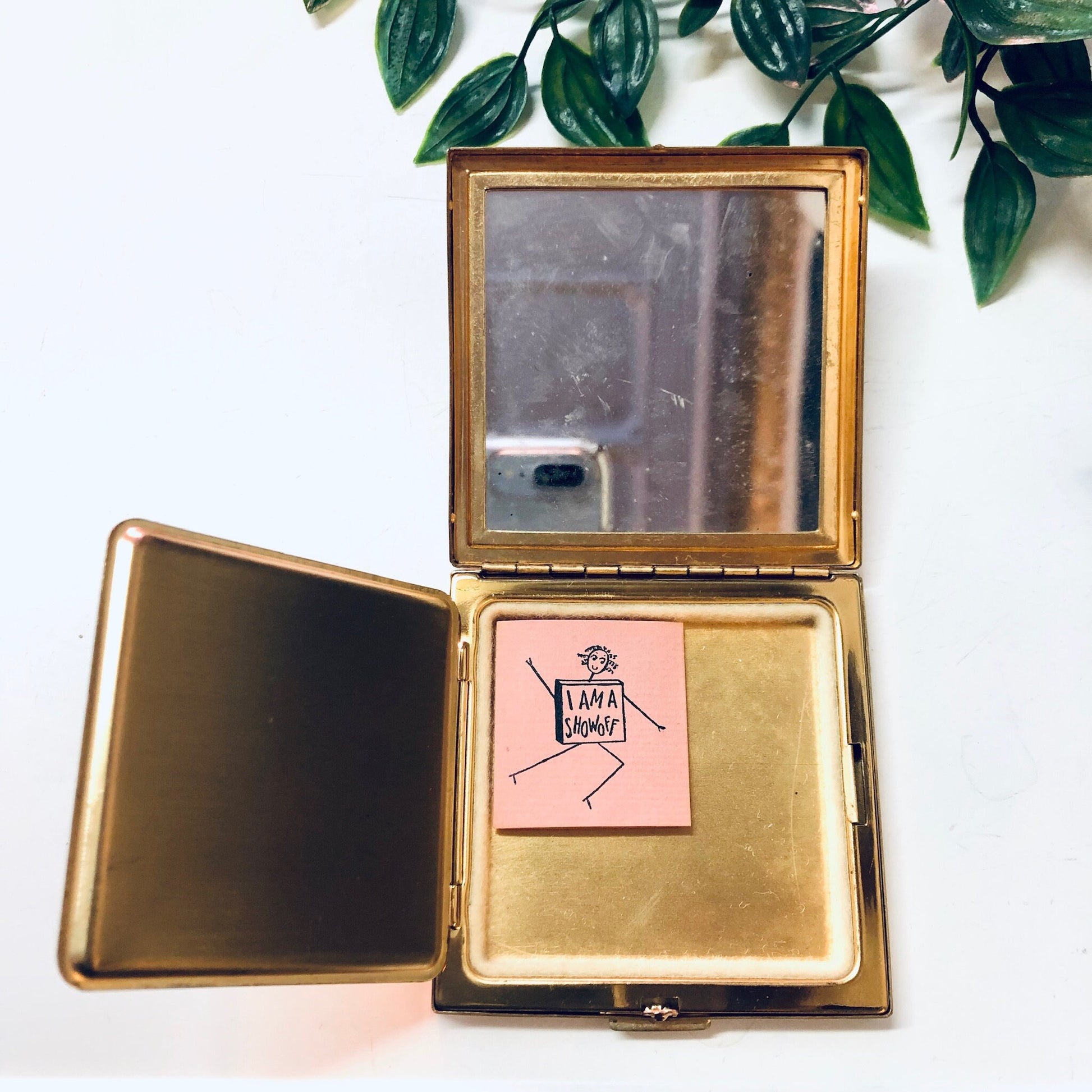 Vintage gold-toned Ciner compact and lipstick case with rhinestones, opened to reveal mirror and pink sticky note, on white background with decorative greenery.