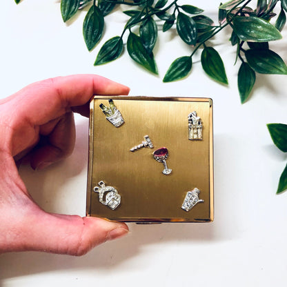 Vintage gold-toned Ciner compact adorned with rhinestone charms including a martini glass, opening to reveal a mirror, held in front of green foliage.