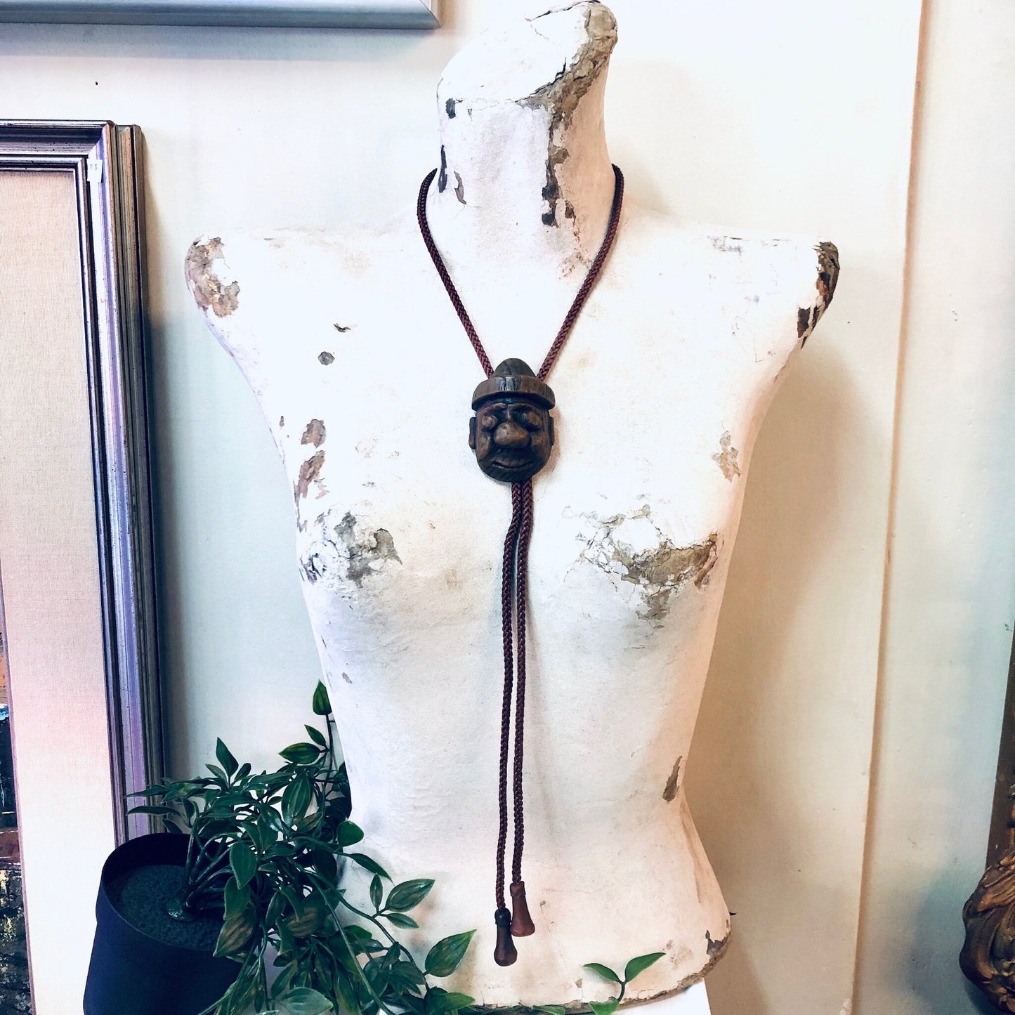 Vintage Japanese bolo tie with carved wooden face pendant on brown rope, mounted on weathered mannequin torso near plants, unique men's accessory.