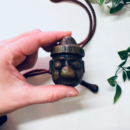 Vintage Japanese wooden carved face bolo tie with brown rope on white background surrounded by green leaves.