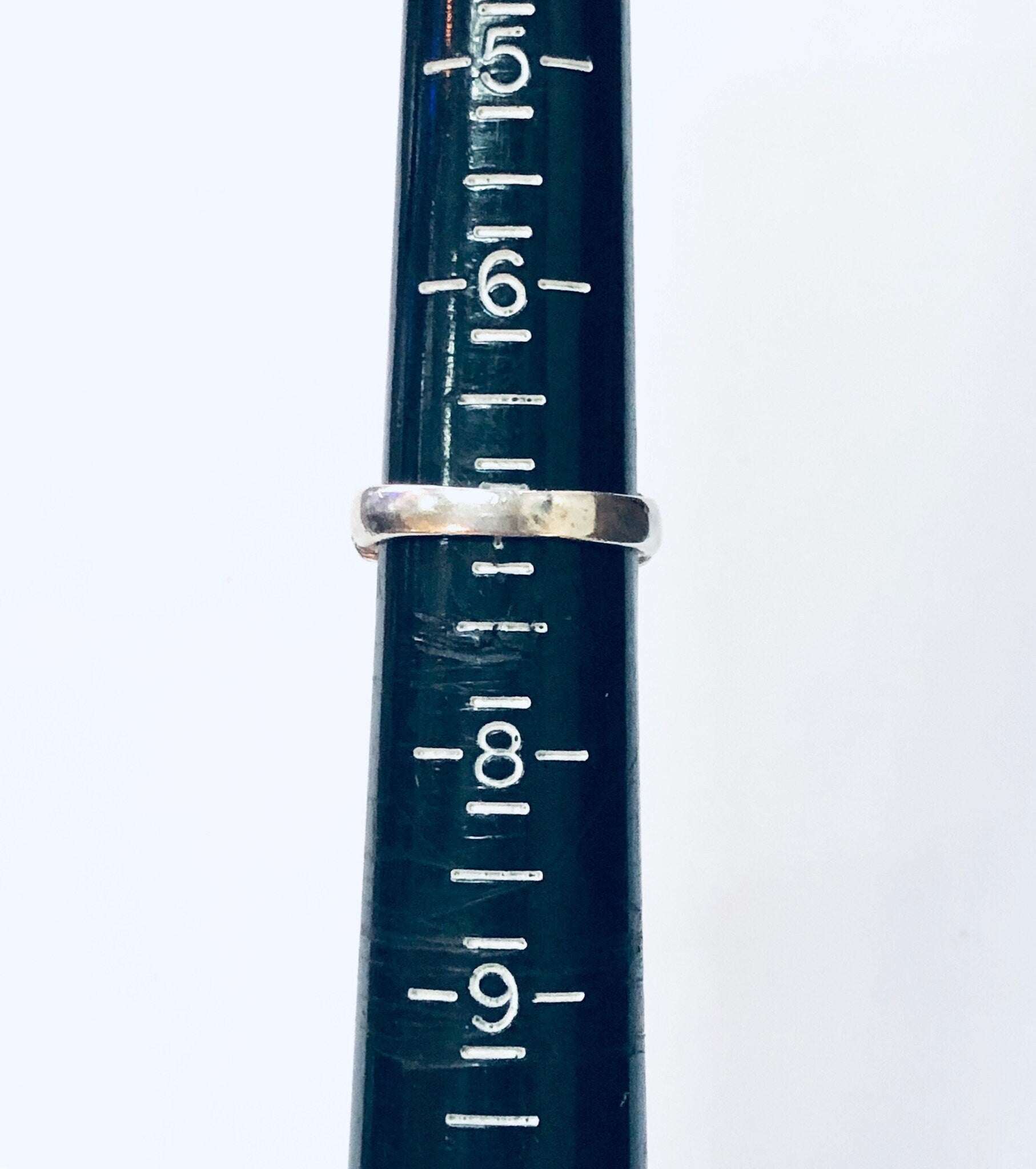 The image shows a close-up view of a vintage silver ring sizer gauge, with markings from sizes 5 to 9 visible on the dark green metal surface.