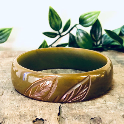 Vintage bakelite bangle bracelet with leaf etching design in green, red and brown colors, placed on weathered wood surface with greenery in background.