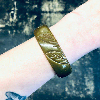 Vintage moss green Bakelite bangle bracelet with etched leaf design, shown on wrist against dark background