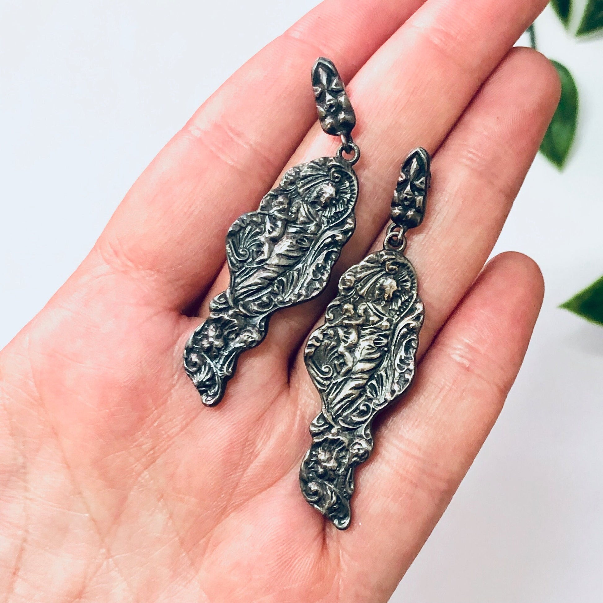 Vintage silver figural earrings in Art Nouveau style featuring detailed lady profile designs, held in a person's hand against a plain background.