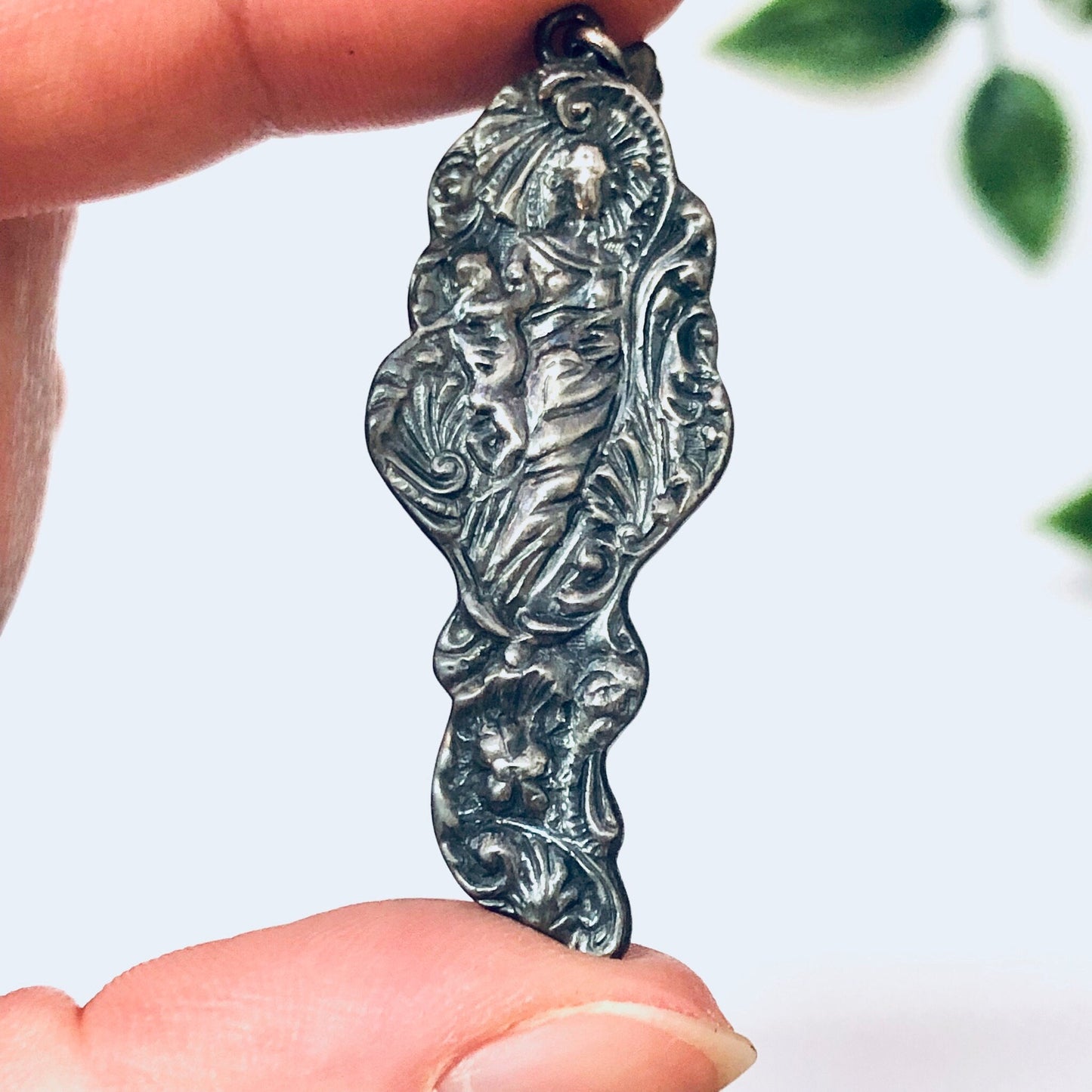 Vintage silver Art Nouveau style dangle earrings featuring an intricate lady figure design, held between fingers against a background of leaves.