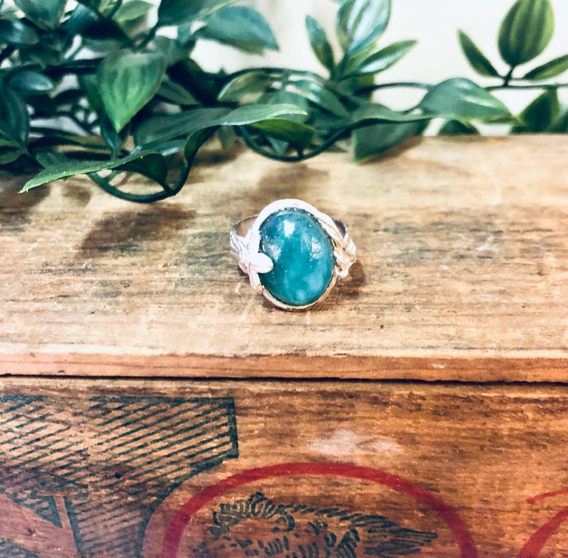 Vintage silver ring with oval green turquoise stone surrounded by floral design on weathered wooden surface with plant leaves in background