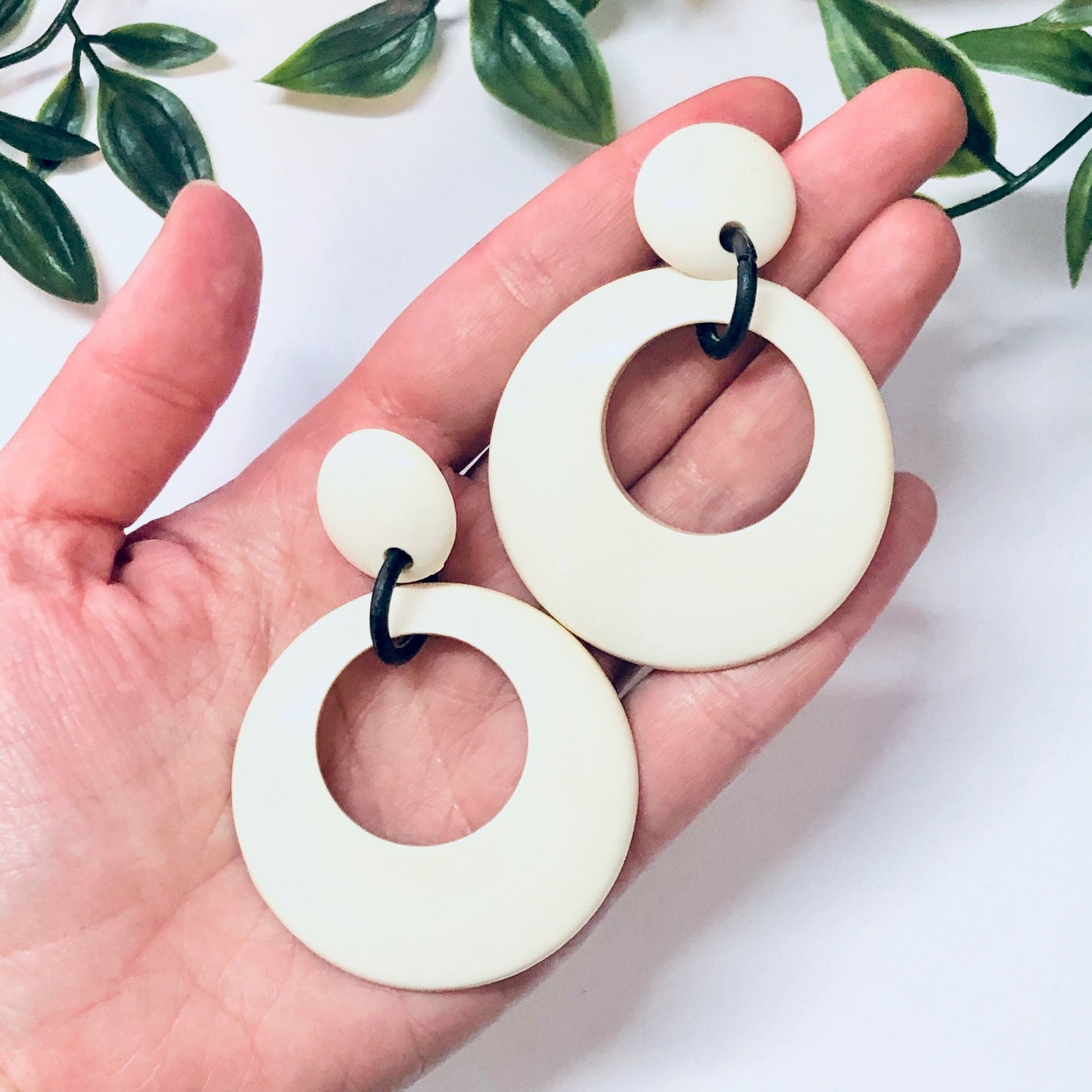 Large white plastic hoop earrings with black accent, held in a hand against a white background with green leaves.