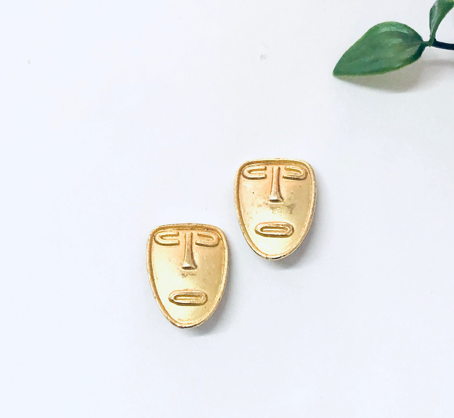 Gold tone abstract face clip-on earrings on white background with green leaf stem