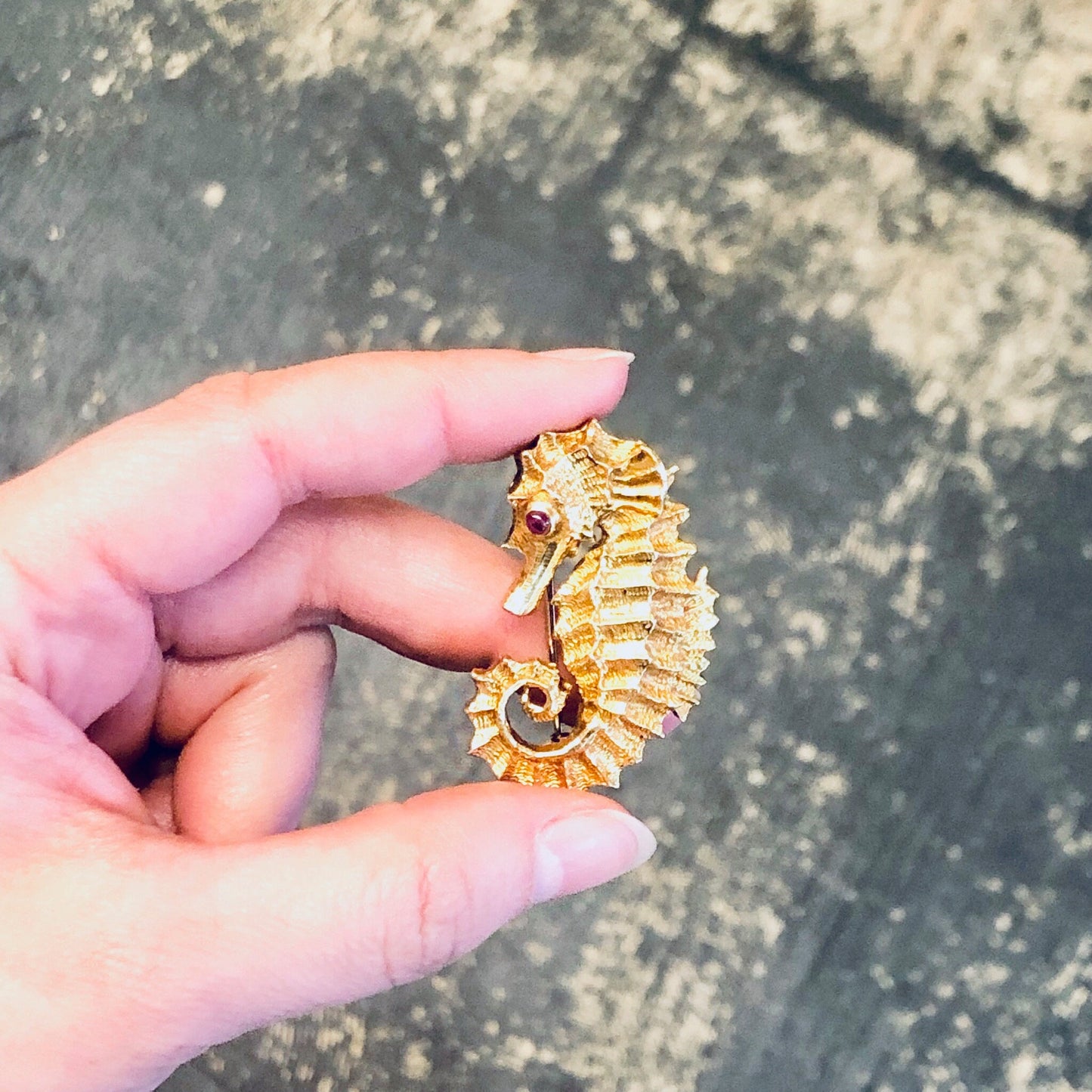 18K yellow gold seahorse brooch with ruby eye, crafted by David Webb in the 1970s, held in a hand against a textured background.