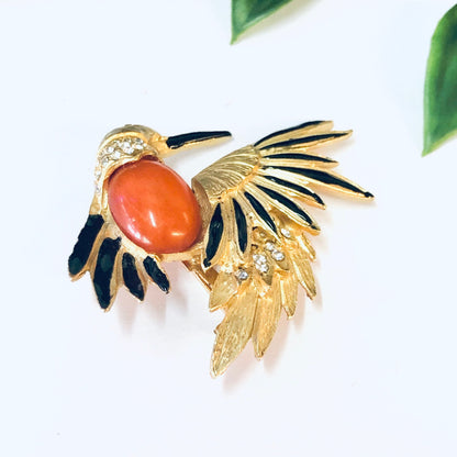 Vintage gold-toned bird brooch pin with orange stone, black enamel accents, and rhinestone details, set against a white background with green leaves.