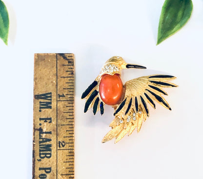 Vintage gold-toned bird brooch pin with black enamel wings, an orange stone body, and rhinestone accents, shown next to leaves and a ruler for scale.