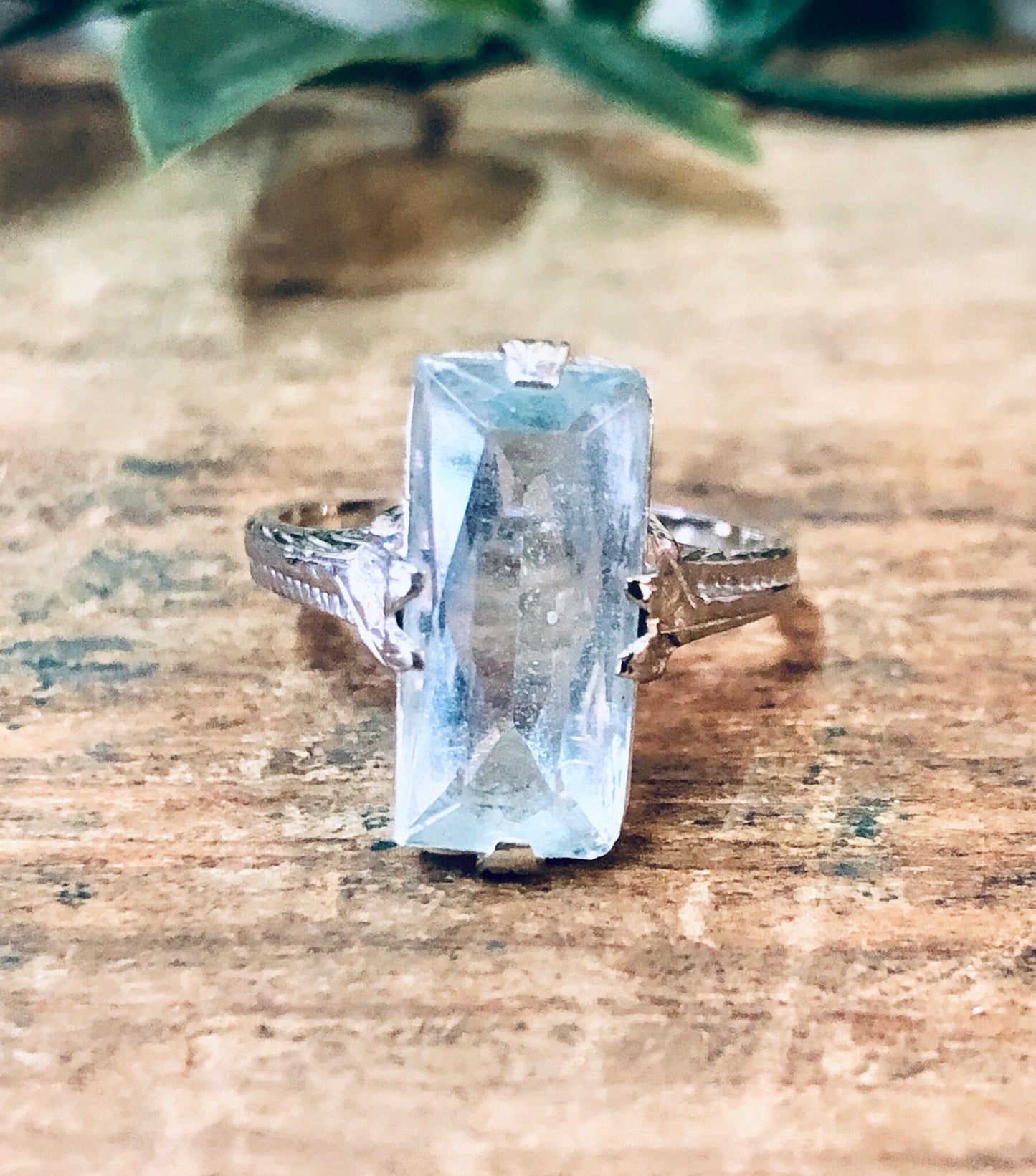 Vintage aquamarine engagement ring in 18K white gold, showcasing an elegant light blue rectangular gemstone on an ornate antique-style setting, photographed on a rustic wooden surface with green foliage in the background.