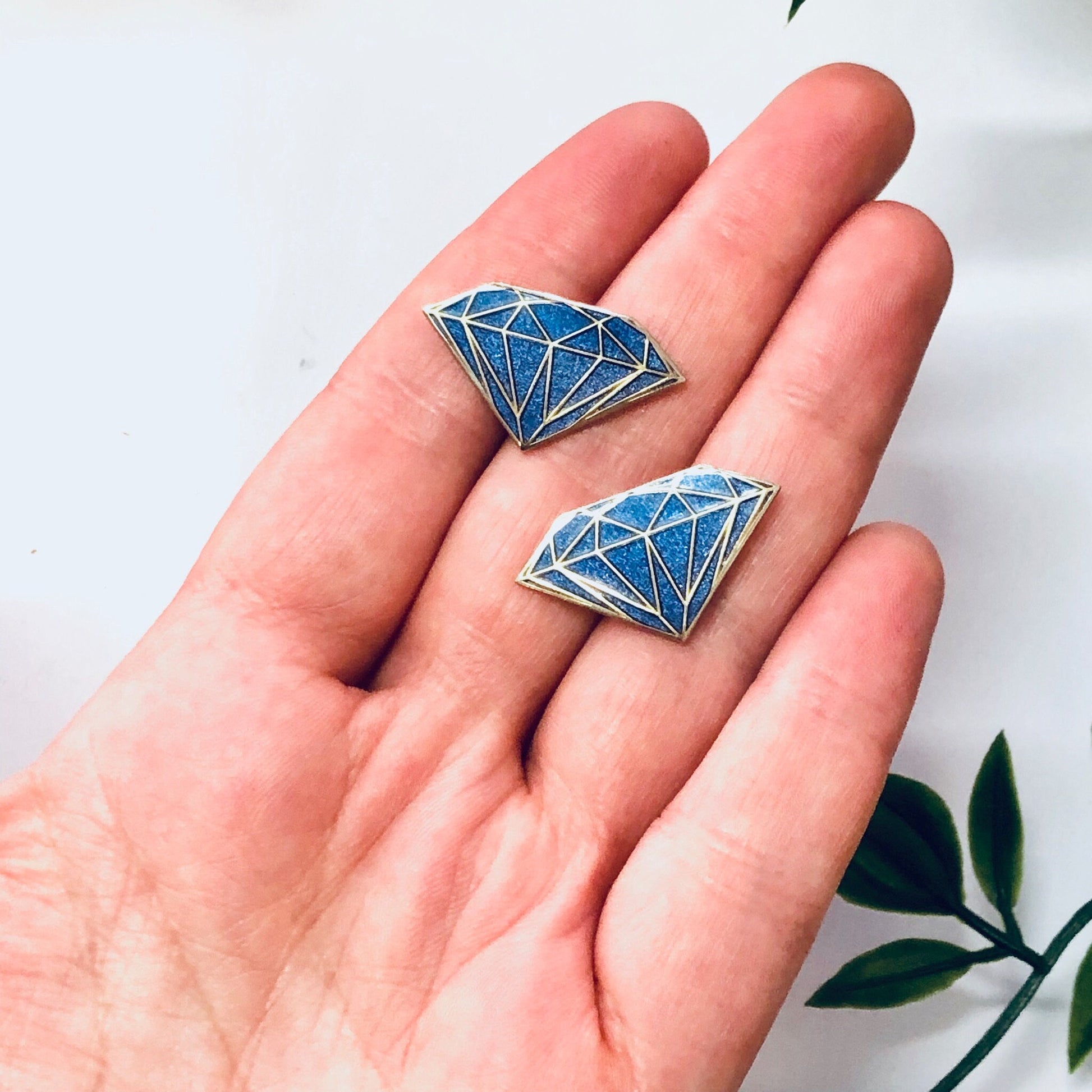 Two blue enamel diamond-shaped earrings held in a person's hand against a white background with leaves.