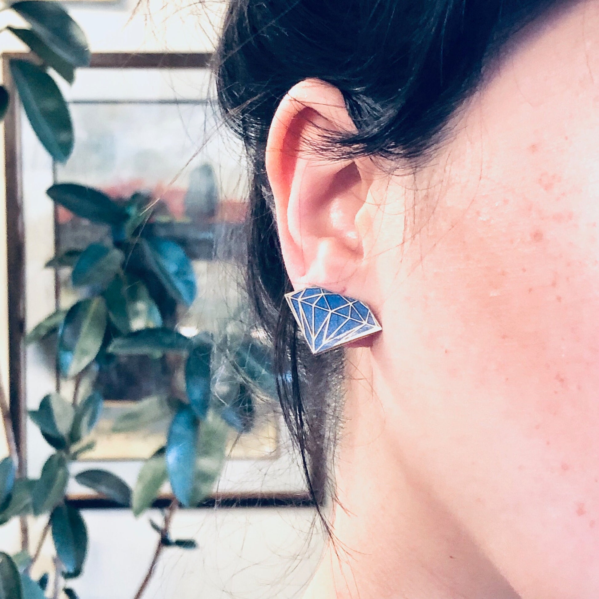 Close-up of a woman's ear pierced with blue diamond-shaped vintage earrings, with plants visible in the background.