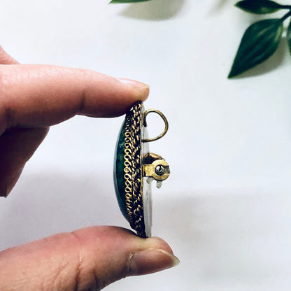 Vintage silver and gold plated brooch pendant with blue and green stones, made in Israel, held between fingers with leaves in background.