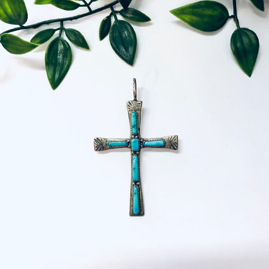 Vintage Zuni silver and turquoise cross pendant surrounded by green leaves on a white background
